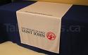 This 30 inch by 60 inch table runner has a logo printed on the front and a web address printed on the top area on a plain white background with blue covered table